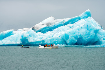 Amphibian vehicle with tourists in Jokulsarlon/Fjallsarlon glacier lagoon by the foot of Vatnajokull volcano. Big blue melting icebergs floats in the lagoon. Overcast day.