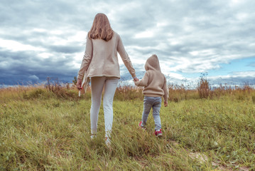 Fototapeta premium Woman mother walks with the child, little son boy 4-5 years old, autumn day outdoors in the park, background field grass and lawn, casual warm clothes with a hood. Back view.