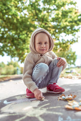 Little boy 3-5 years old, happy draws colorful crayons on pavement, creativity in park, feel happy. Casual warm clothes with hood. Autumn day. Emotions happiness and pleasure.
