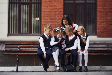 School kids in uniform that sits outdoors on the bench with smartphone