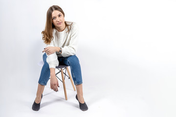 Fototapeta premium attractive young girl in jeans and a white sweater sits on a chair on a white background, copy space