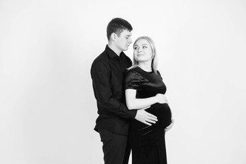 Pregnant woman and her husband hugging and holding onto your stomach in black clothes on a white background. Black and white picture. Beautiful married couple. Copy, empty space for text
