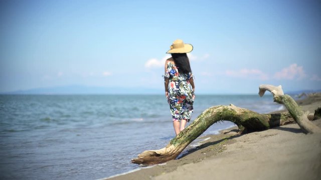 Romantic woman is walking on the beach with colorful long skirt swaying in the wind dress. Waves of the sea are hitting carrying slippers in hand woman's feet. Alone barefoot mossy dry tree trunk bark