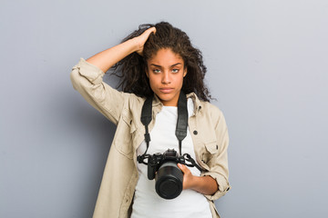 Young african american photographer woman holding a camera being shocked, she has remembered...