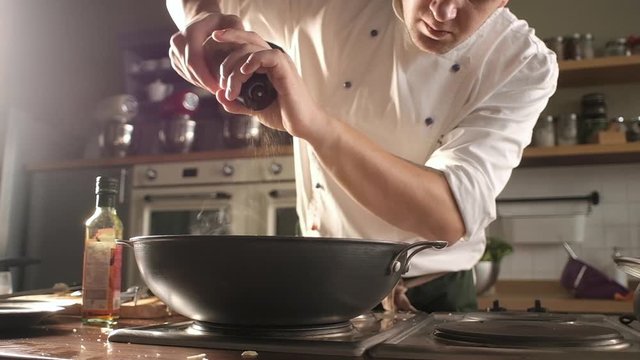Cooking delicious dish in modern kitchen. the chef in uniform pours pepper from the pepper bowl on the prepared dish. A wok or frying pan is on the stove.  Delicious dinner at home or in a restaurant