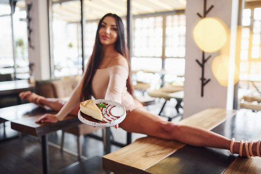 Young brunette in sexy wear soing stretches by sitting on the table and holds plate with food in restaurant