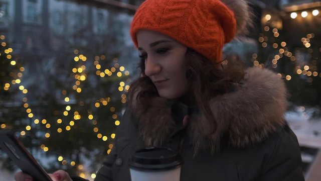 Portrait of an attractive girl in a bright hat with a phone and coffee in winter on a background of lights