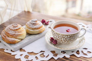 A cup of tea with roses buds, sweet buns with sugar powder on handmade white napkin and wooden table near window with day light. Beautiful breakfast or dessert time.