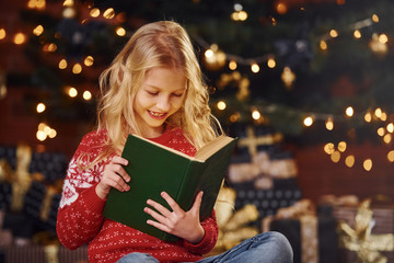 Cute little girl in red festive sweater reading book indoors at christmas holiday time