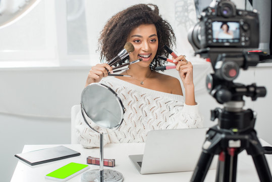 selective focus of happy african american influencer in braces holding lip glosses and cosmetic brushes near smartphone with green screen and digital camera - Powered by Adobe