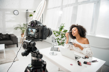 selective focus of curly african american influencer in braces applying makeup near digital camera