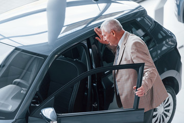 Aged man in formal wear walking in the automobile salon and looking for the car
