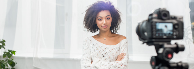 panoramic shot of curly african american influencer standing with crossed arms and looking at...