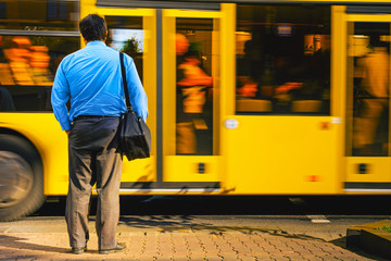 Fototapeta na wymiar A man in a blue shirt in the bustle of the city against the backdrop of a passing bus. Man near the road view from the back. Tired man returns from office work. Work routine concept.