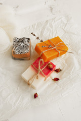 Beautifully packaged natural colorful soap with thin rope, natural loofah, dried rose buds and craft white paper on a light concrete background. Natural self care concept. Top view at an angle