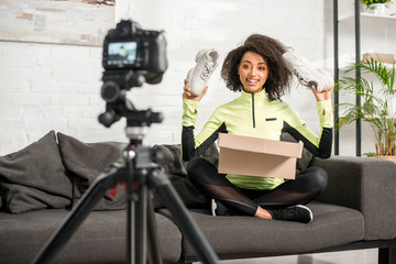 Fototapeta na wymiar selective focus of happy and sportive african american influencer in braces holding new sneakers near box and digital camera