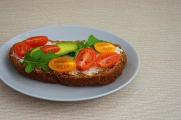 Sandwich with fresh vegetables arugula, avocado and tomatoes on bread with cheese in a white plate on a beige background