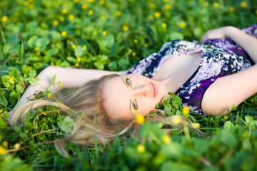 Portrait of young beautiful blond woman lying in grass and flowers on summer day with green nature landscape