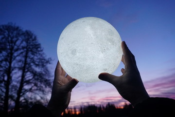 Round full moon in hands against evening sky. Lunar model, moon-shaped lamp with moon craters