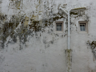 Grungy once white wall with dirt and mildew with two small windows and a drain pipe