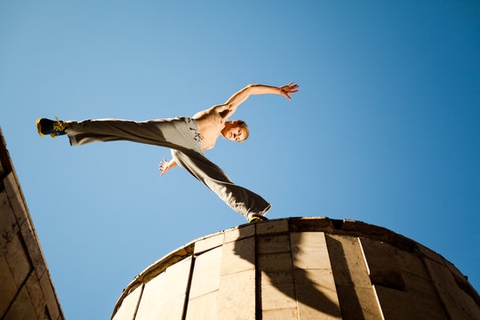 Young man jumping and practicing parkour between buildings roofs outside on clear summer day with blue sky at background