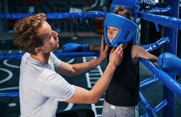 Young boxing coach is helping little boy in protective wear on the ring between the rounds