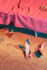 Pink vintage ladies shoes and cocktail glass in sand next to sunbed with pink towel and ashtray on it.