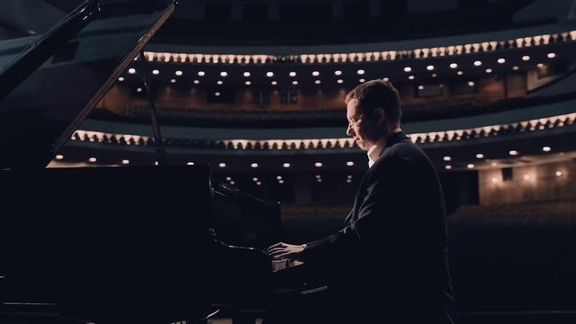 Handsome middle-aged pianist playing on a grand piano on big stage in concert hall with dimmed light 