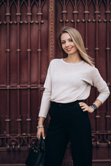 Outdoor photo of blonde lady posing on architecture background in autumn day.Close up fashion street style portrait. wearing cute trendy outfit.Dark casual trousers and creamy sweater.Fashion concept.