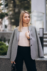 Outdoor photo of blonde lady posing on architecture background in autumn day.Close up fashion street style portrait.wearing dark casual trousers,creamy sweater and gray coat or jacket.Fashion concept.