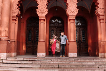 Fototapeta na wymiar Young stylish pretty couple of hipsters in love are having fun in the city near castle in spring. young blond woman in red dress and man with beard are smiling on a date