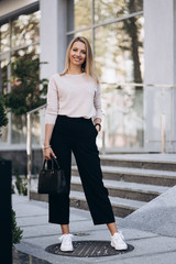 Outdoor photo of blonde lady posing on architecture background in autumn day.Close up fashion street style portrait. wearing cute trendy outfit.Dark casual trousers and creamy sweater.Fashion concept.
