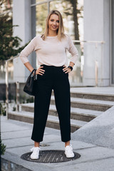 Outdoor photo of blonde lady posing on architecture background in autumn day.Close up fashion street style portrait. wearing cute trendy outfit.Dark casual trousers and creamy sweater.Fashion concept.