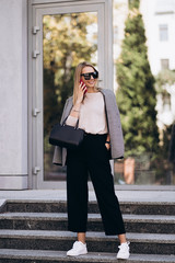 Beautiful blonde woman talking on phone walking on street.Portrait of stylish smiling business woman in Dark casual trousers and creamy sweater.Fashion concept.. Female business style.High Resolution.
