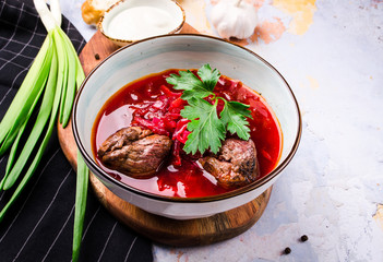 Traditional Ukrainian Russian Borscht with bacon, hot soup with beets, cabbage and carrots in a bowl on a wooden board. Served with green onions, sour cream, rye bread, garlic, greenery.