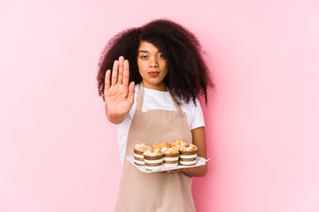 Young afro pastry maker woman holding a cupcakes isolatedYoung afro baker woman standing with outstretched hand showing stop sign, preventing you.