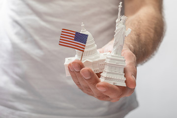 cropped view of man holding small statue of liberty and united states capitol near little american flag isolated on white - Powered by Adobe