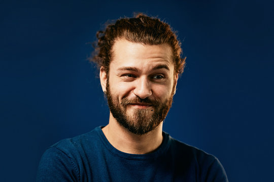 Young attractive caucasian bearded hipster with curly hair dressed in sweater standing in front of blue background with one eyebrow raised and looking away.