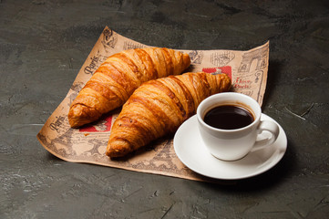 Croissants and a cup of coffee with saucer on craft paper on a dark background, vintage style
