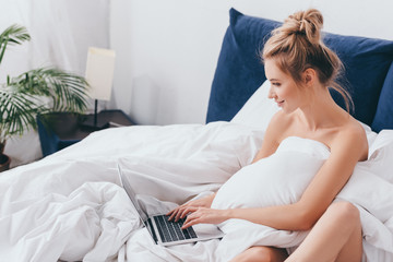 smiling woman using laptop on bed with sheets in morning