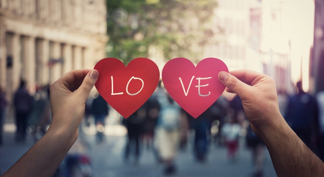 Two hands holding different paper heart symbols, unite them to create the word Love, on a crowded street. Woman and man togetherness concept. Valentine's day holiday background, romantic composition.
