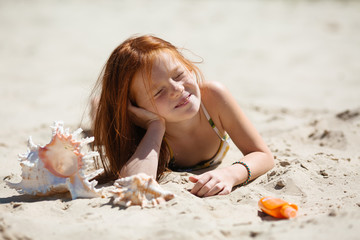 little long-haired red-haired girl with her eyes closed lies on a sandy beach with her face exposed to the sun