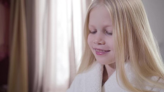 Close-up of blond Caucasian girl opening red lipstick and shaking head. Stylish pretty kid in white bathrobe choosing cosmetics at home. Beauty, care, lifestyle.