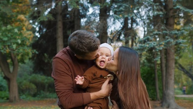 Young mother and father holding cute little child in their hands and kissing him from both sides outdoors. Cheerful child is smiling. Happy family having fun in park.
