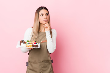 Young baker woman holding sweets looking sideways with doubtful and skeptical expression.
