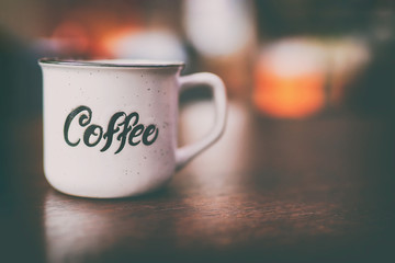 Enamel white metal mug with  inscription  coffee on  wooden table on blurred background. Place for text or advertising