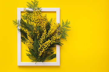Mimosa flowers and green branches  on yellow pattern texture of crumpled paper with white photo frame, yellow background. Spring and Easter Frame Concept, flat lay, blank space