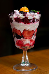 Colorful trifle dessert with fizalis, strawberries, blueberries, jam and whipped cream on high translucent glass on wooden table, vertical photography in low-key lightning