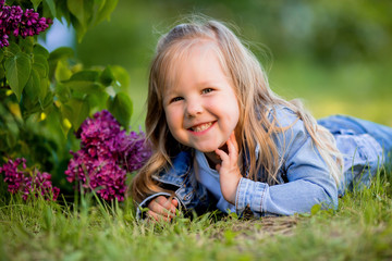 little blonde girl lies near the lilac flowers on the green grass and smiles