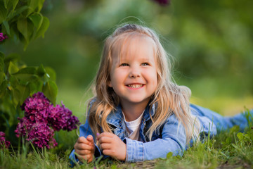 little blonde girl lies near the lilac flowers on the green grass and smiles